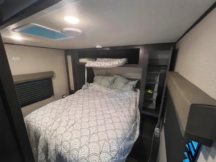 RV rental's cozy bedroom: main bed, upper bunk, window with blind, and built-in storage shelves.. Grand Design Transcend Xplor 2022