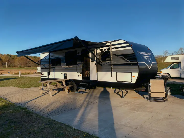 Transcend XPLOR travel trailer RV rental at a sunny campsite with awning extended. Dog peeks from the doorway.. Grand Design Transcend Xplor 2022