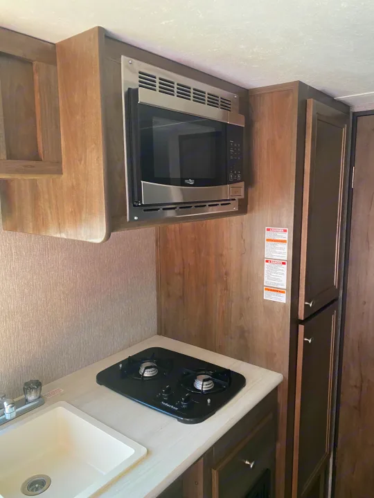 Cozy RV rental kitchen with an overhead microwave, two-burner stovetop, and sink. Wood cabinets and textured walls complete the space.. Dutchmen Coleman Lantern LT 2020