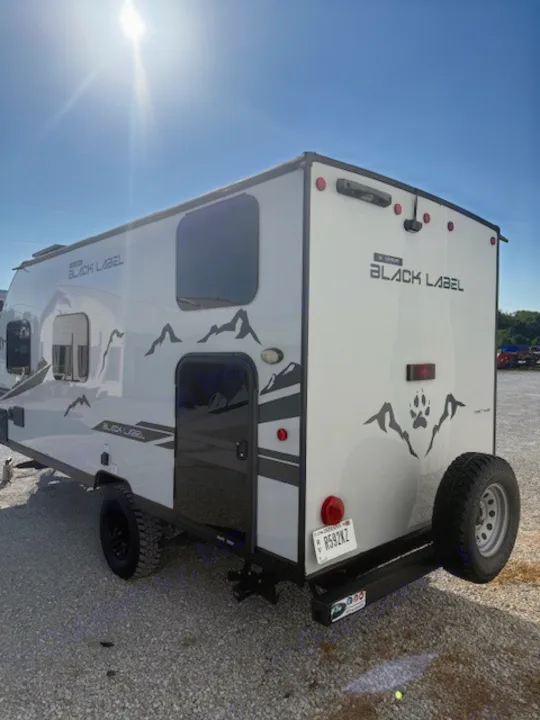 A sleek white Black Label travel trailer RV rental, featuring mountain graphics and a rear-mounted spare tire, parked on gravel.. Cherokee WOLF PUP  16BHSBL 2023