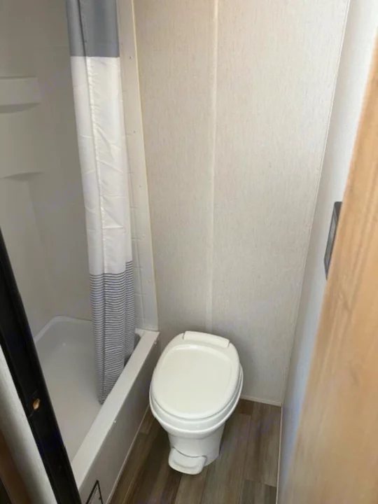 Compact RV rental bathroom. Features a white shower/tub with striped curtain (left) and a white toilet (right) on wood-look floor.. Cherokee WOLF PUP  16BHSBL 2023