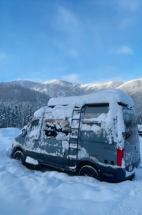 A gray Storyteller Overland van, suitable for an rv rental adventure, is heavily snowed in with deep drifts & snowy mountains behind it.. Storyteller Overland Storyteller Overland Class B 2021