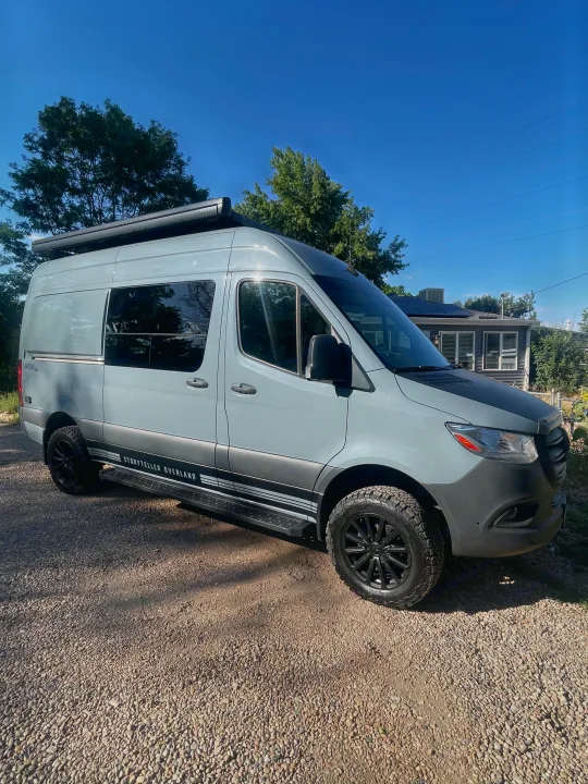 A modern gray Storyteller Overland adventure van with black wheels, parked on gravel. Consider this RV rental for your next trip.. Storyteller Overland Storyteller Overland Class B 2021