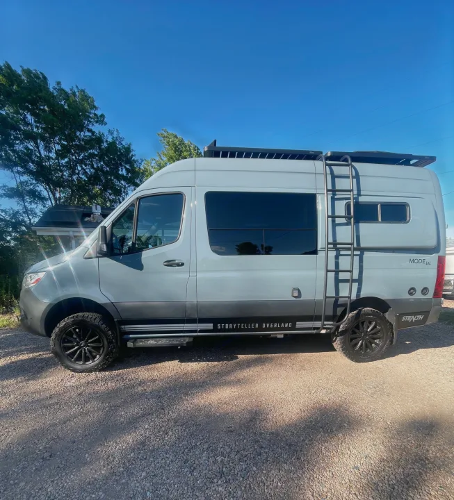 This gray Storyteller Overland Mode 4x4 camper van, ideal for rv rental, is parked on gravel with blue sky & trees.. Storyteller Overland Storyteller Overland Class B 2021