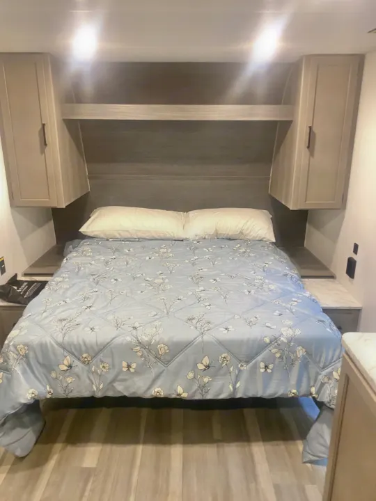 Compact RV rental bedroom with light walls, a bed with blue floral comforter, overhead cabinets, and side shelves.. Keystone RV Hideout Sport 2024