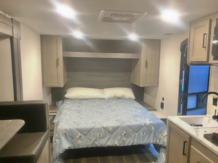 Cozy RV rental interior with a light blue floral bed, overhead storage, and integrated kitchen/dinette space.. Keystone RV Hideout Sport 2024