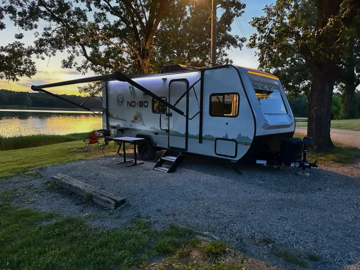 Serene lakeside RV rental setup at sunset. Travel trailer with extended awning, outdoor table, and grill. Perfect for camping.. No Boundaries 19 2021