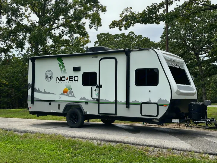 A white No Boundaries travel trailer, perfect as an RV rental, is parked on an asphalt patch surrounded by green trees.. No Boundaries 19 2021