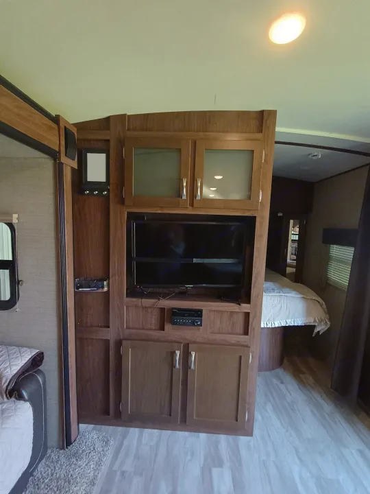 Spacious RV rental interior featuring a built-in wooden entertainment center with TV, storage, and light-colored flooring, looking towards the bedroom.. Dutchmen Aerolite 28dbhs 2017