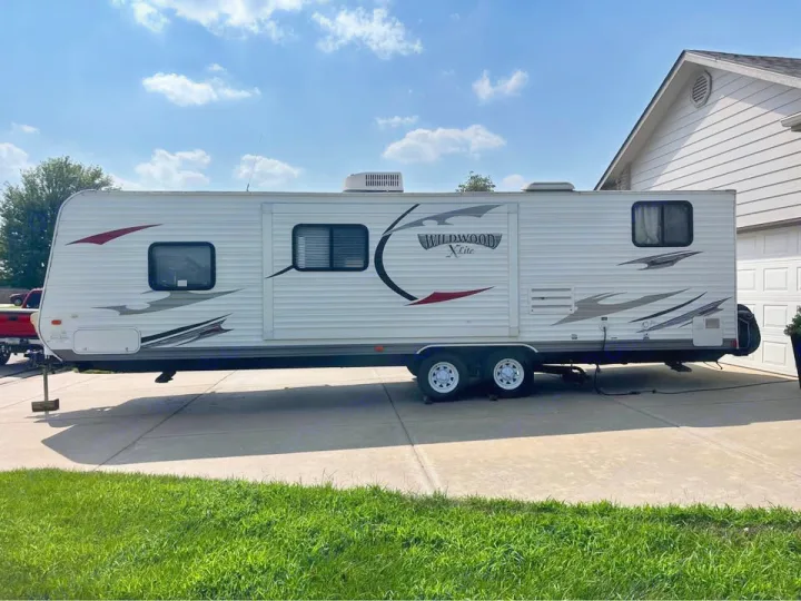 White Wildwood X-Lite travel trailer RV rental, parked on a paved driveway next to a house on a sunny day.. Forest River Wildwood X-Lite 2013