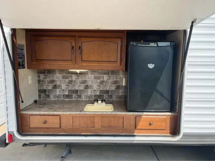 Exterior RV rental kitchen: wooden cabinets, tiled backsplash, sink, mini-fridge, and counter for camping.. Forest River Wildwood X-Lite 2013