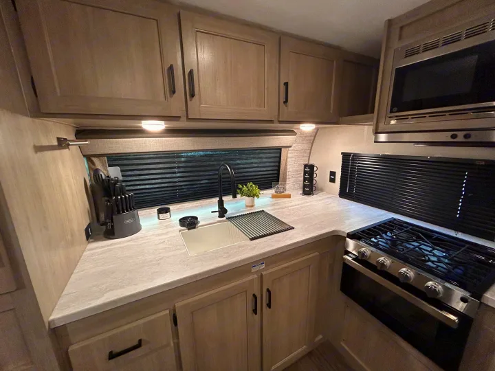 Step inside this RV rental kitchen, featuring modern light wood cabinets, dark appliances, a sink, and a stove.. Forest River 26FKDS 2025