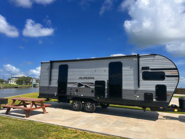 This Aurora RV rental, a grey travel trailer, sits at a waterfront campsite with a picnic table under blue skies.. Forest River 26FKDS 2025