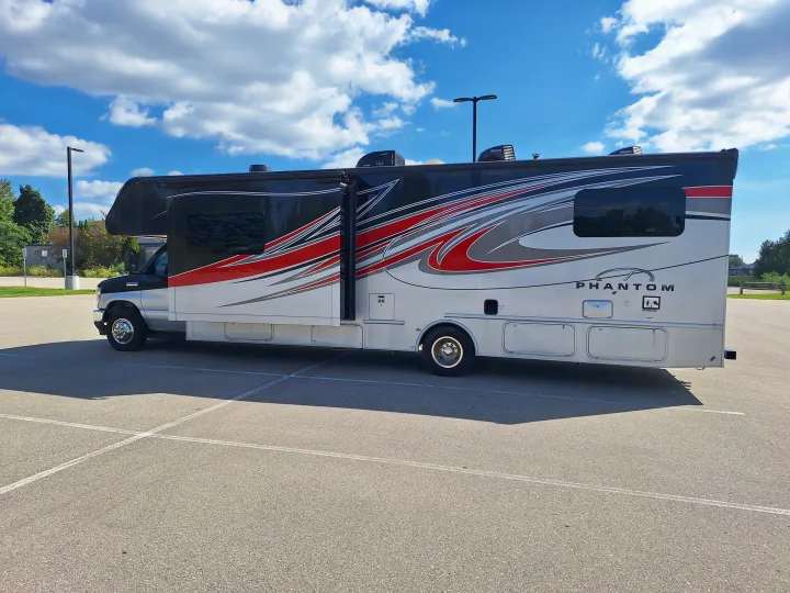 A white, black, and red Class C RV rental is parked with its slide-out extended, under a bright, cloudy sky.. Nexus Phantom 2024