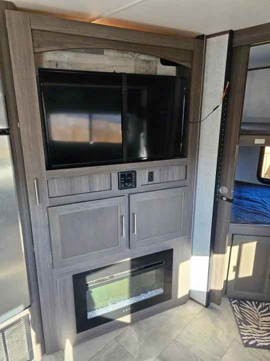 Modern RV rental interior: TV, electric fireplace, and cabinets in entertainment center. Refrigerator and bunk beds are also visible.. Dutchmen Kodiak Ultra Lite 2021