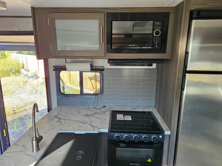 Inside an RV rental: a modern kitchen with a sink, microwave, stove, and fridge, set against a gray tiled backsplash.. Dutchmen Kodiak Ultra Lite 2021