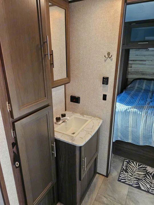 This RV rental interior shows a compact bathroom with a sink, mirror, storage, and wall controls, next to a bedroom.. Dutchmen Kodiak Ultra Lite 2021