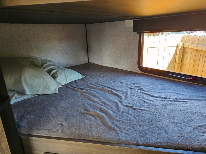 An RV rental's cozy bunk bed, featuring soft gray bedding, green pillows, and a window view of a wooden fence.. Dutchmen Kodiak Ultra Lite 2021