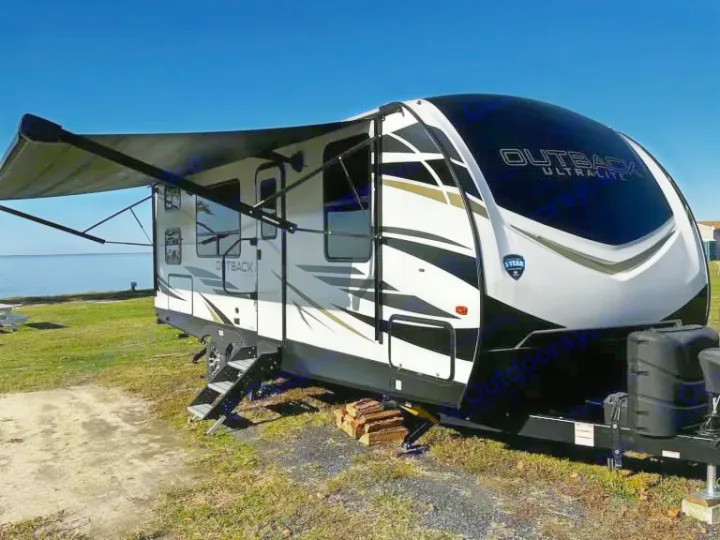 Waterfront RV rental: A sleek Outback Ultralite travel trailer with an extended awning parked by the clear blue water.. Keystone RV Outback Ultra Lite 2024