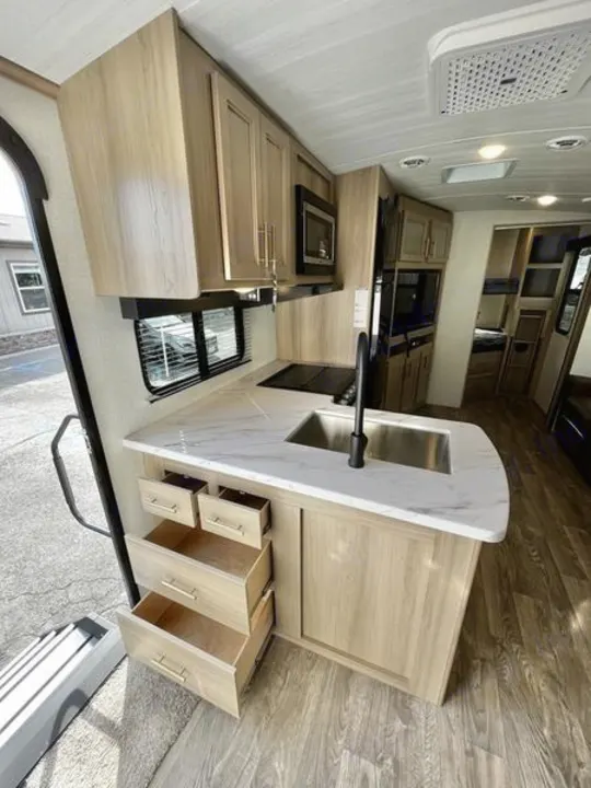 Modern RV rental interior: well-equipped kitchen with light wood cabinets, sink, and appliances, ready for adventure.. Keystone RV Outback Ultra Lite 2024