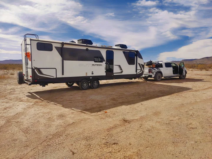 White RV travel trailer, branded 'Outback', hitched to a pickup truck, parked in a sunny, arid landscape. Ideal for rv rental.. Keystone RV Outback Ultra Lite 2024