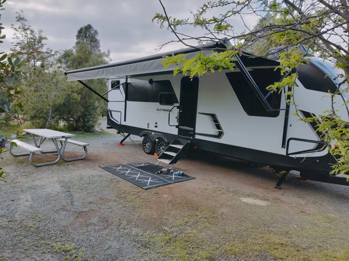 RV rental campsite setup: modern travel trailer with extended awning, picnic table, and a pet-friendly mat.. Keystone RV Outback Ultra Lite 2024