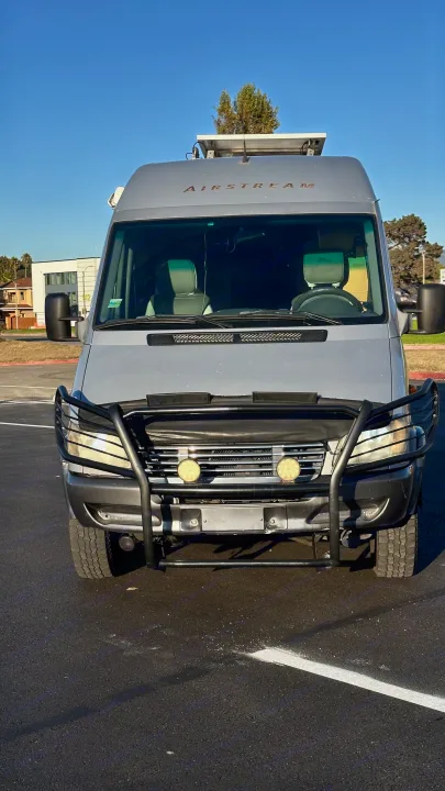 This grey Airstream van, an rv rental, showcases a front bull bar with auxiliary lights and roof-mounted solar panels.. Airstream Interatate 2003