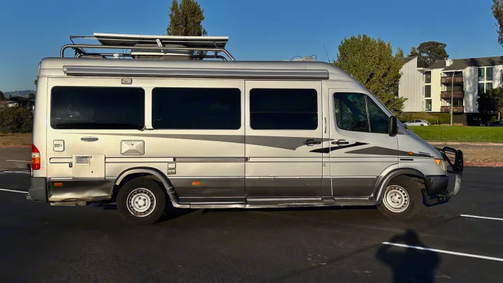 Side view of a silver Mercedes Sprinter RV rental, featuring tinted windows and roof rack, parked on asphalt. Ready for travel.. Airstream Interatate 2003