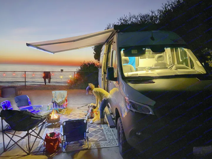 An RV rental campervan lights up a beach evening with a person, dog, and warm campfire at sunset.. Winnebago Revel 2024
