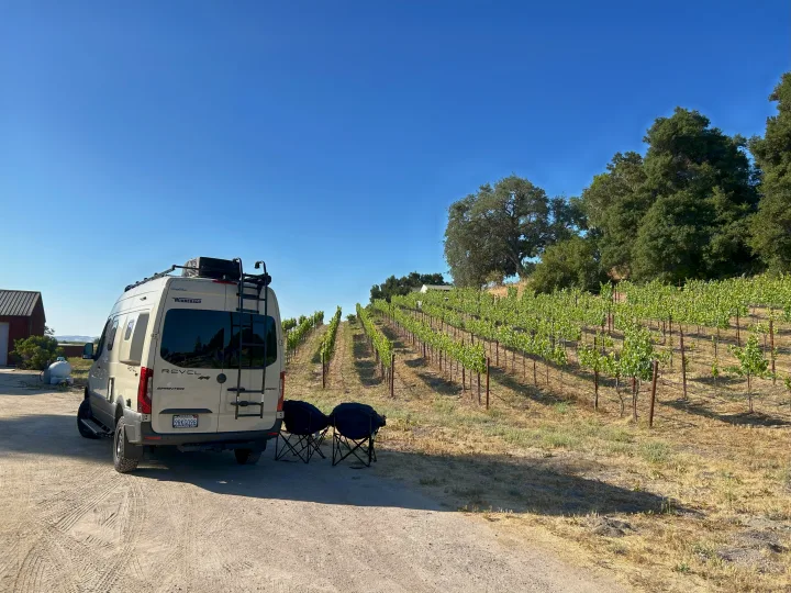 Scenic RV rental adventure: A white Winnebago Revel van is parked by lush vineyard rows, with chairs set for relaxation.. Winnebago Revel 2024
