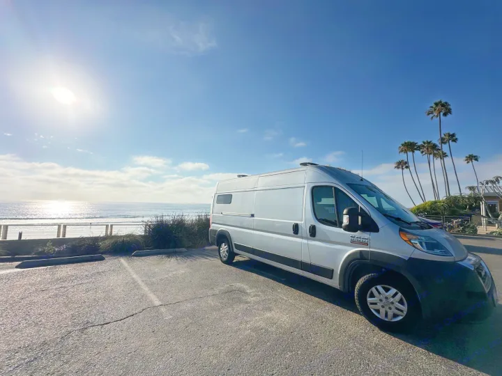 Sunny day! A silver RV rental van is parked in a beachside lot overlooking the ocean and palm trees.. Dodge Ram ProMaster 2021