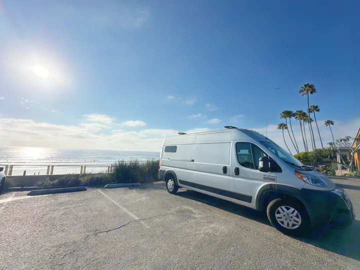 Silver RV rental van parked on a sunny day by the ocean, with surfers in waves and tall palm trees along the coast.. Dodge Ram ProMaster 2021
