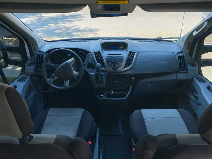 Interior of an RV rental: a Ford van dashboard, steering wheel, media controls, and front passenger seats are visible.. Ford Transit 2018