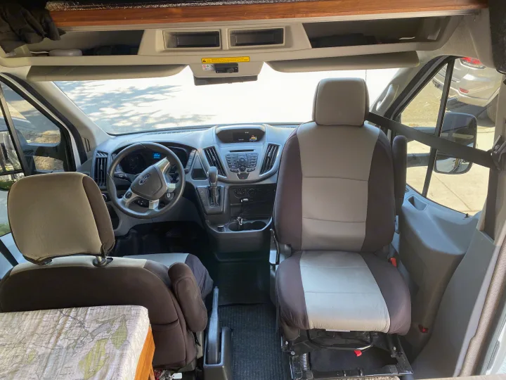 Inside this RV rental: front cabin view showing comfortable driver & passenger seats, dashboard, and overhead storage.. Ford Transit 2018