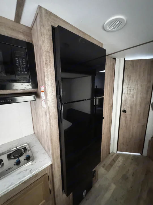 Modern RV rental kitchen featuring sleek black appliances: microwave, gas cooktop, and large reflective refrigerator built into wood-look cabinetry.. Gulf Stream Enlighten 2025