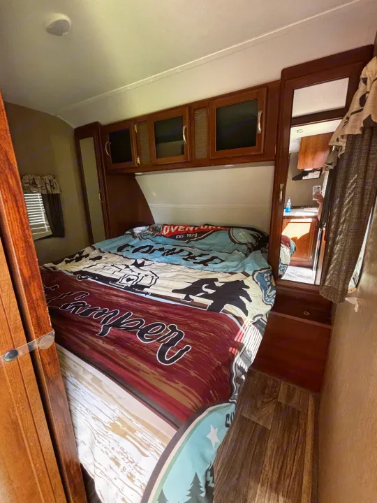 Cozy RV rental bedroom, featuring a large bed with a 'Camper Adventure' comforter, storage, and a kitchen reflection.. Keystone RV Passport Grand Touring 2016