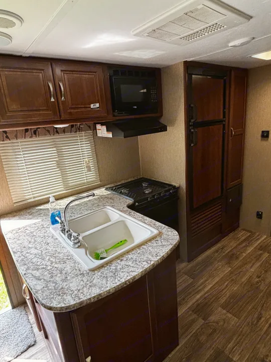 RV rental kitchen features a double sink, 2-burner stove, microwave, fridge, wood cabinets, granite-look counters, and ceiling AC.. Keystone RV Passport Grand Touring 2016