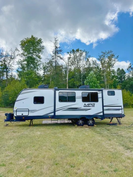 A white MPG RV rental travel trailer set up in a grassy field, surrounded by a lush forest under a mix of clouds and blue sky.. Cruiser RV MPG Ultra Lite 2022
