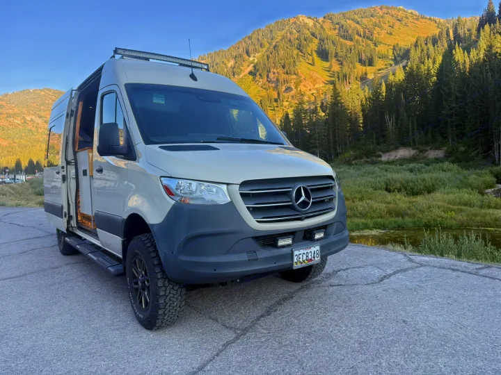 Adventure-ready beige Mercedes-Benz Sprinter camper van, possibly an rv rental, by mountains and water. Ideal for off-grid trips.. Mercedes-Benz Sprinter 2019