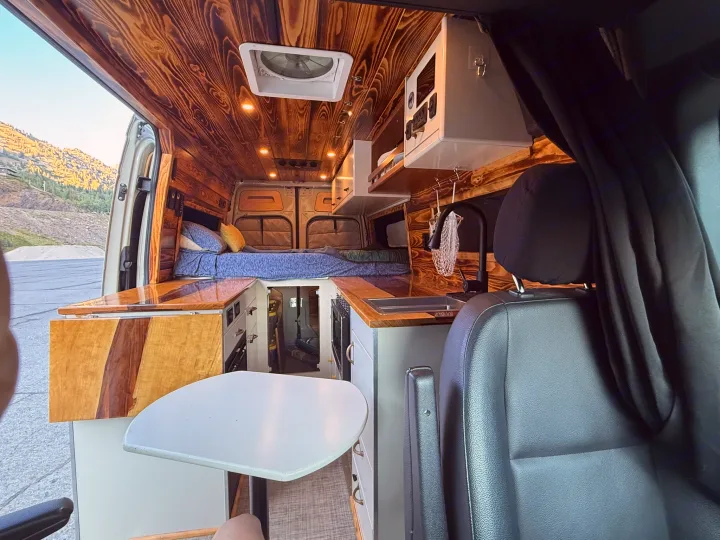 Interior of a cozy RV rental showcasing a wood-paneled ceiling, comfortable bed, compact kitchen, and dining table.. Mercedes-Benz Sprinter 2019