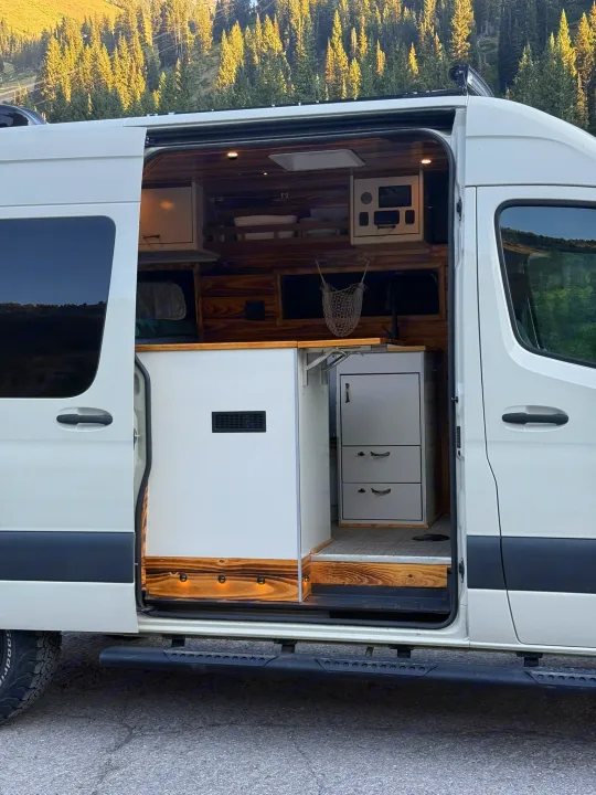 White RV rental camper van with open door, revealing custom wood interior, kitchen, and control panel. Perfect for adventure.. Mercedes-Benz Sprinter 2019
