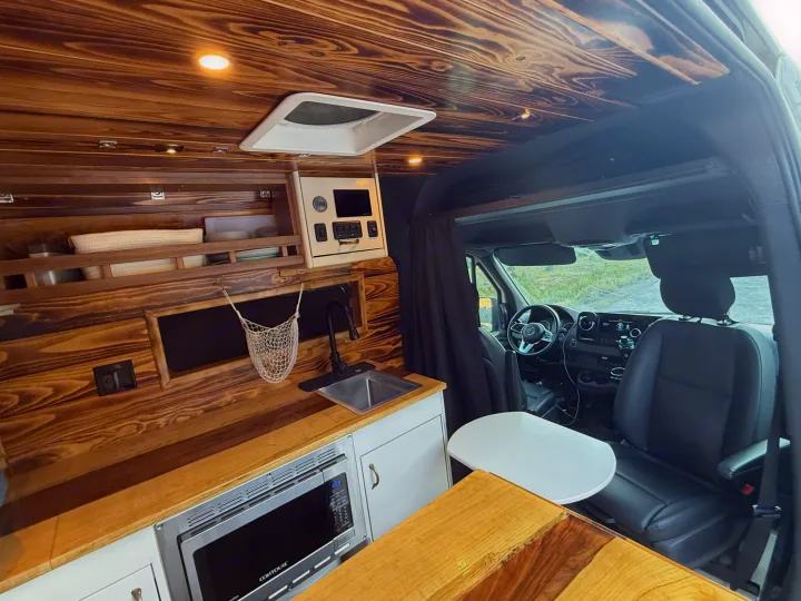 Custom wood-paneled RV rental interior, featuring a compact kitchen with microwave, sink, and comfortable driver's cab.. Mercedes-Benz Sprinter 2019