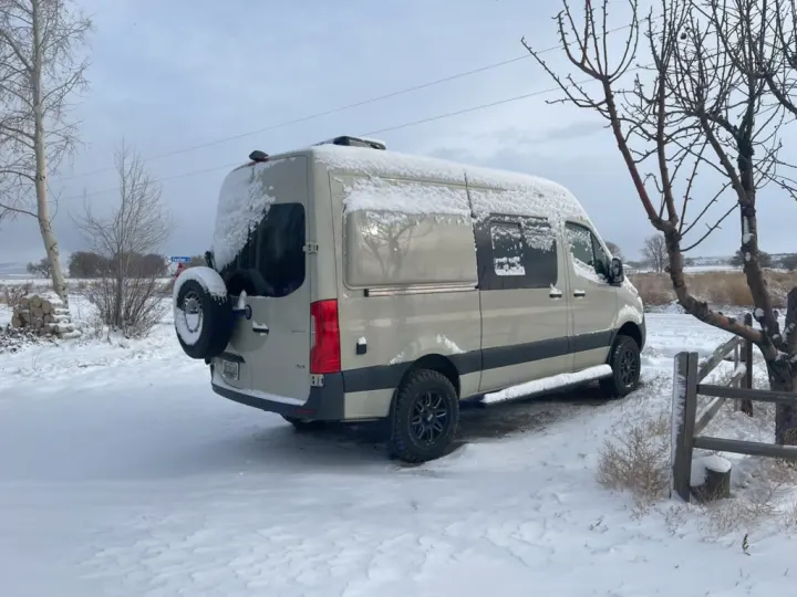A light-colored camper van, an RV rental, dusted with snow in a wintry landscape. Perfect for snowy travels & winter camping.. Mercedes-Benz Sprinter 2019