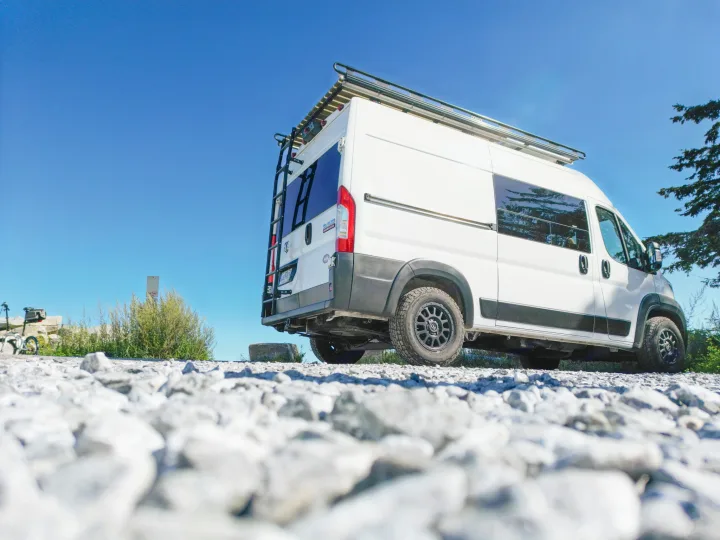 A white adventure camper van, equipped for off-road travels, awaits its next rv rental journey on a sunny day. Low angle view on gravel.. Dodge Ram Promaster 2500 2020