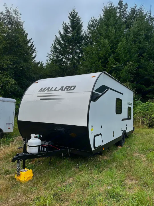 White Mallard travel trailer rv rental parked in a grassy area amidst tall green trees. Perfect for your next adventure.. Heartland Mallard 2025