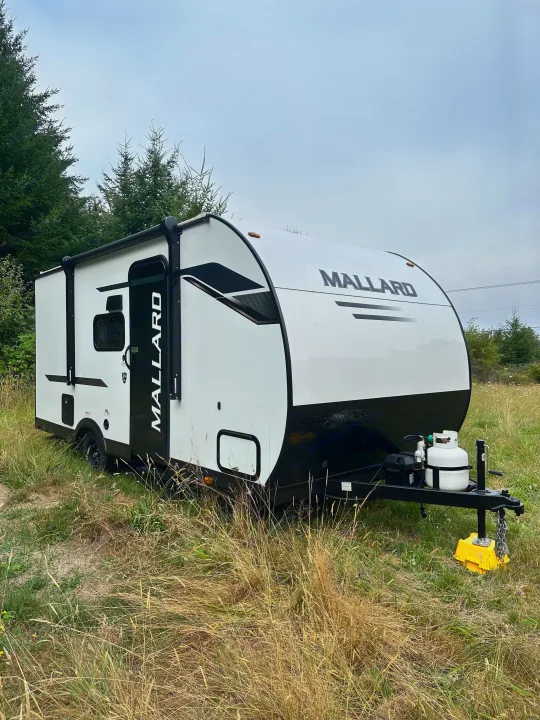 This white and black Mallard RV rental, a travel trailer, is parked in a grassy field with trees under a cloudy sky.. Heartland Mallard 2025