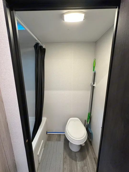 Inside an RV rental: a bright, compact bathroom featuring a toilet, shower with black curtain, and wall-mounted mop.. Heartland Mallard 2025