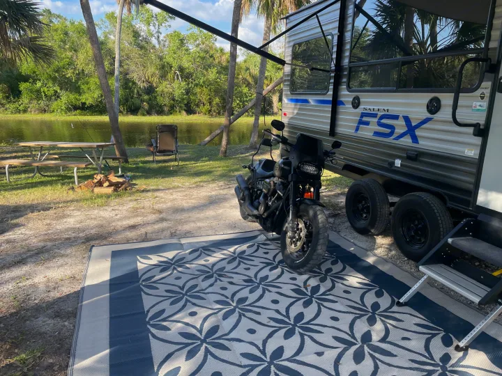 RV rental at a serene lakeside campsite with a motorcycle, picnic table, and outdoor rug setup.. Forest River Salem 2021