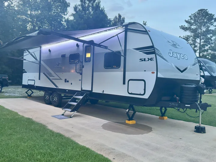 An RV rental, a Jayco travel trailer, is brightly lit by its awning lights at dusk, parked on a concrete pad.. Jayco Jay Jay Flight 294QBS 2025