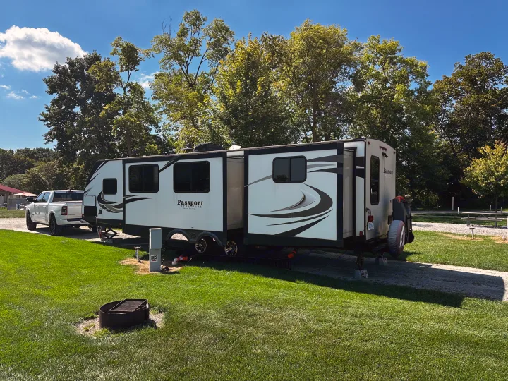 A white travel trailer, likely an RV rental, extends its slides at a sunny green campsite with a truck and fire pit.. Keystone RV Passport Grand Touring 2017